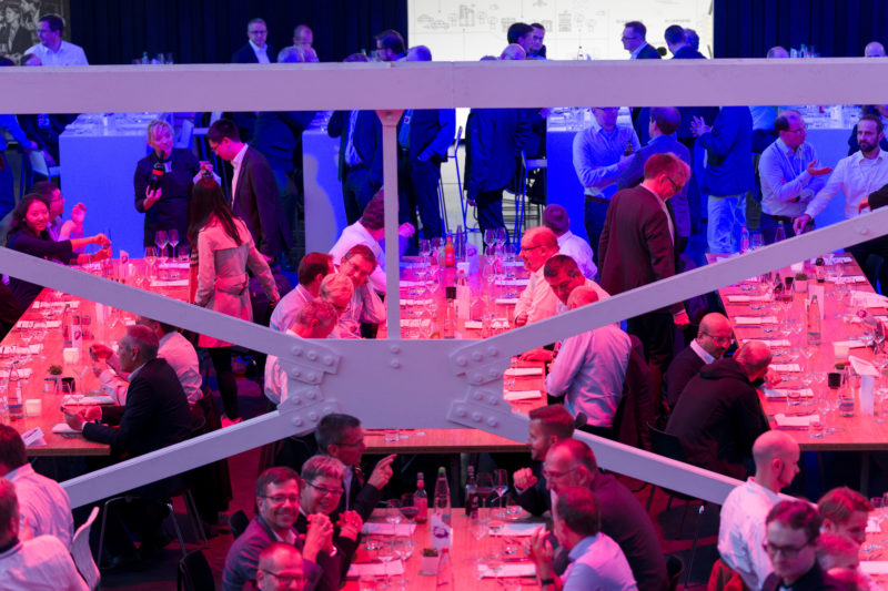Editorial photography as event photography and fair photography: During the evening program of a company event, the participants sit opposite each other at long tables before dinner and talk excitedly. The former factory hall is bathed in blue and red light. Other participants prefer to stand with their colleagues in the corridor.