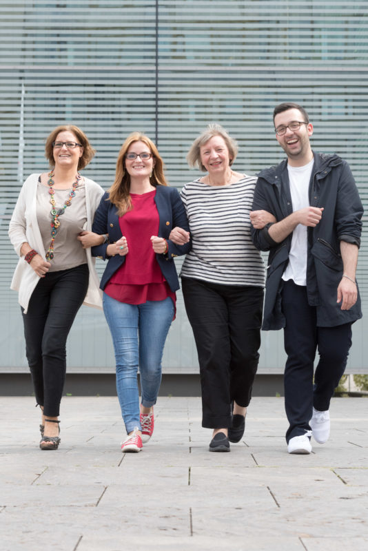 Group photo: Four municipal employees are working together on a project. They walk towards the camera with their arms hooked on, laughing.