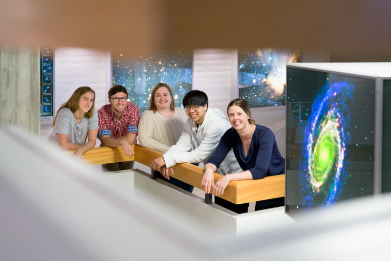 Gruppenfoto: Gruppenportrait einer Arbeitsgruppe in der Astronomieforschung. Alle stehen im Treppenhaus und sind von Fotos von Sternengalaxien umgeben.