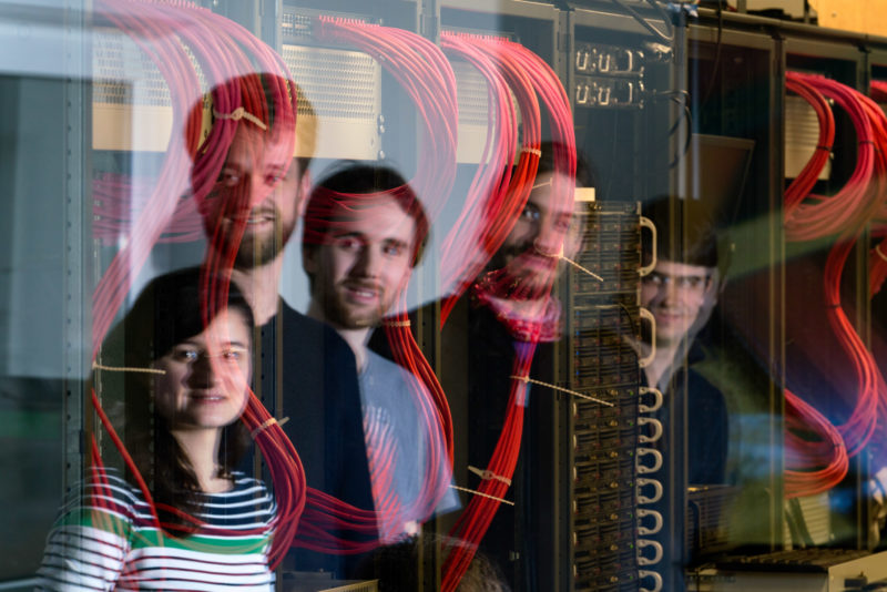 Group shot: Portrait of a working group at a computing institute of a university. The employees are reflected in the glass pane to the server room, where thick red cables lead into the computer cabinets.