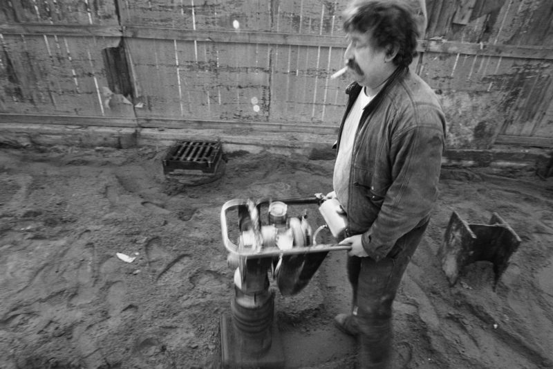 Schwarz-Weiss-Reportagefotografie an verschiedenen Arbeitsplätzen Anfang der neunziger Jahre: Ein Arbeiter im Straßenbau bewegt mit einer Zigarette im Mund einen Rüttler über eine Sandschicht, die dann später zugepflastert werden soll.