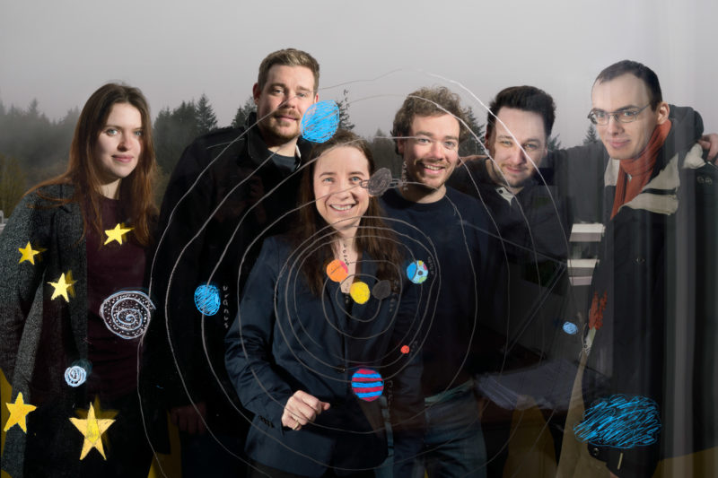 Group shot: Six colleagues from an astronomy research group stand behind a glass pane on which the child of an employee has painted the Earth