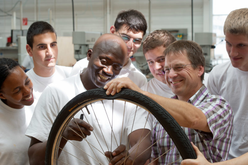 Reportagefotografie: Im Rahmen der betrieblichen Ausbildung reparieren Auszubildende in ihrer Fahrradwerkstatt Diensträder. Ein junger Flüchtling steht in der Mitte der Gruppe, während der Ausbilder erklärt, wie man einen Reifen wechselt.