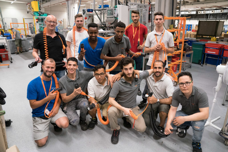 Group photo: Trainers and trainees at a cable manufacturer. The trainees come from many countries, there are some refugees with them. All of them have charging cables for electric vehicles in their hands or have them hanging around their neck.