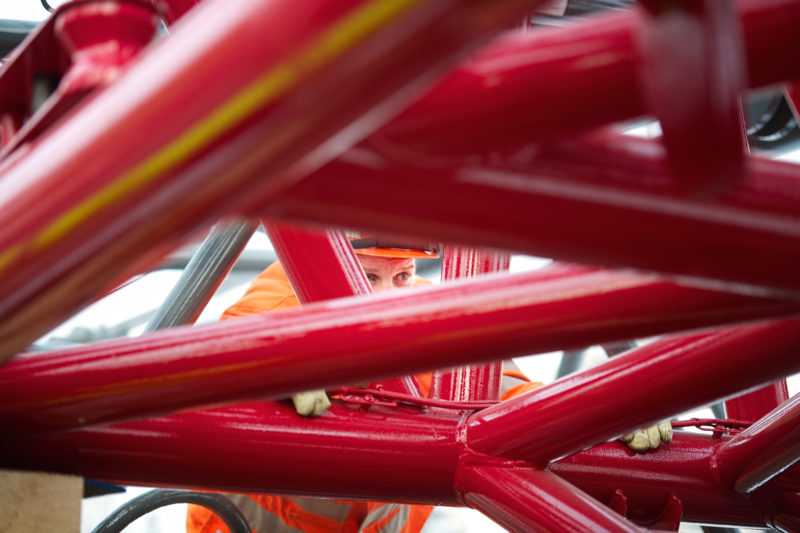 Industriefotografie: Ein Mitarbeiter eines Logistikunternehmens für den Güterverkehr auf Schienen schaut durch ein kompliziertes rotes Gestänge eines Krans, der auf einen Güterwagen gesetzt wird, um dann an seinen Bestimmungsort in Osteuropa transportiert zu werden.