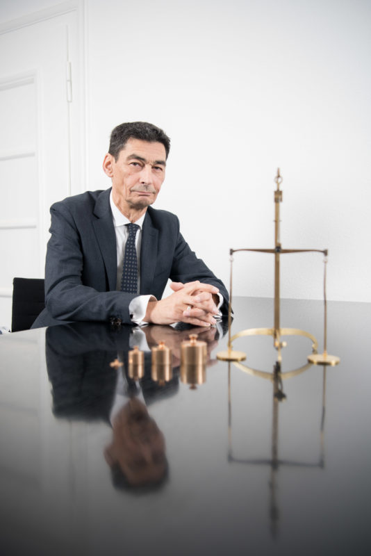 Managerportrait: Ein Bankmanager sitzt mit gefalteten Händen an einem spiegelnden schwarzen Tisch.Vor ihm steht eine goldene mechanische Waage mit nebeneinander aufgereihten Gewichten. Das Foto ist mit Blitzanlage ausgeleuchtet und wird für die Öffentlichkeitsarbeit verwendet.