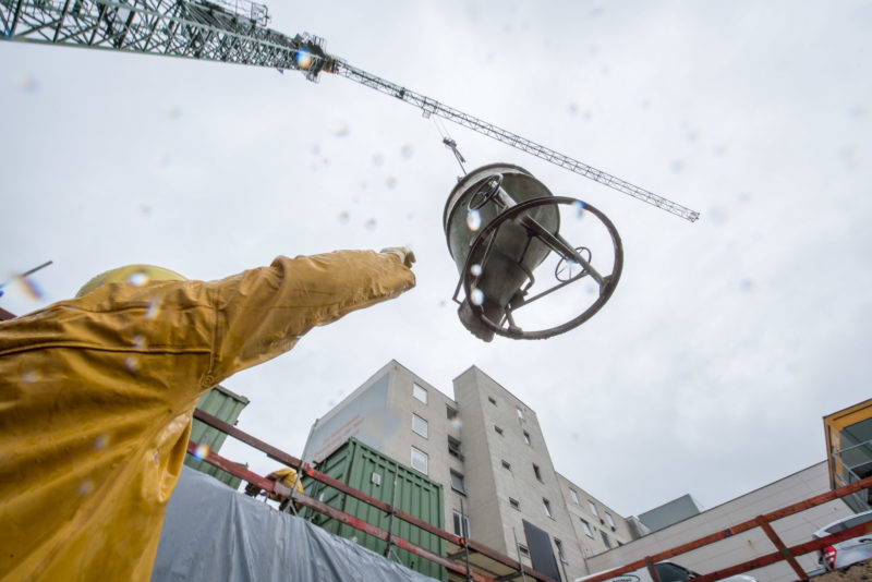 Editorial photography: In the rain a full concrete bucket floats on the crane to the construction site. A worker reaches for it to empty it at the right place.