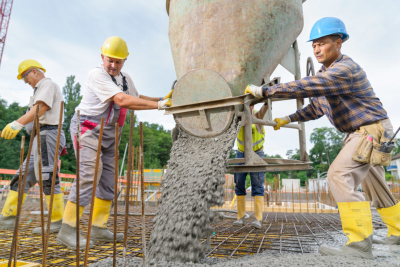 Reportagefotografie: Bauarbeiter positionieren einen am Kran hängenden Betonkübel, aus dem sich der Beton in die eisenarmierte Verschalung der Zwischendecke ergießt.