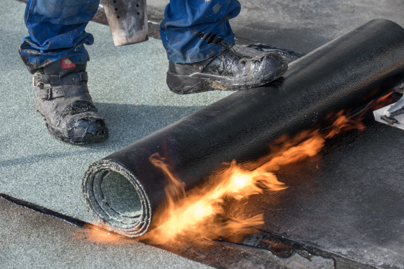 Reportagefotografie: Ein Dachdecker rollt mit den Füßen Bitumen-Dachpappe aus, während er sie mit einem Flammenwerfer verklebt.