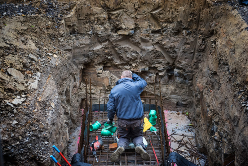 Reportagefotografie: Ein Arbeiter kniet in einer Erdgrube um Drainage-Rohre zu verlegen. Man sieht ihn von hinten.