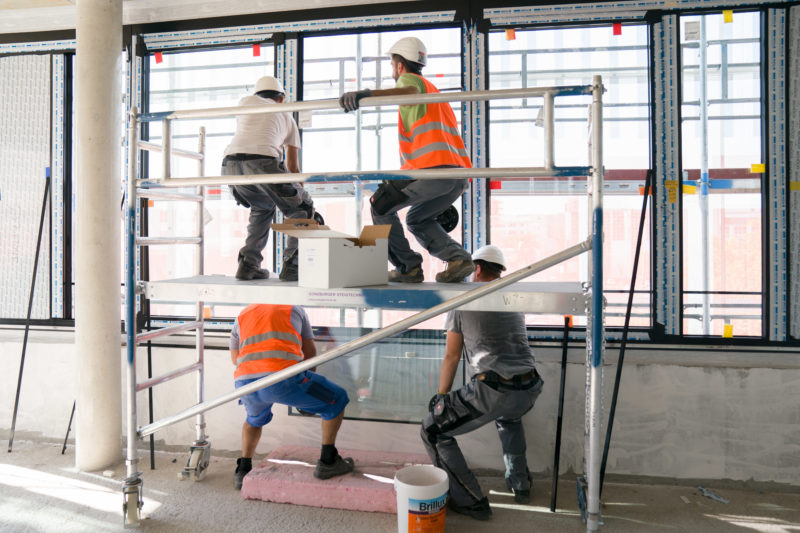 Editorial photography: Four window manufacturers lift a large glass pane into position in the shell of an office complex. Two stand on the floor and two on a scaffolding.