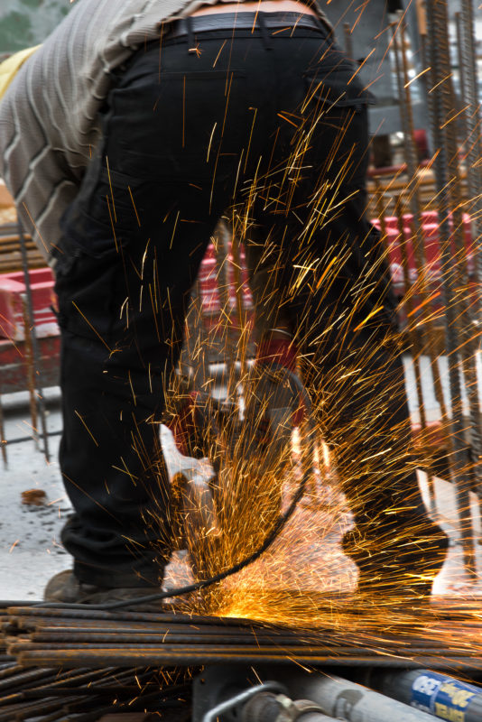 Editorial photography: An iron braider saws reinforcing steel. Orange sparks spray away from the cutting.