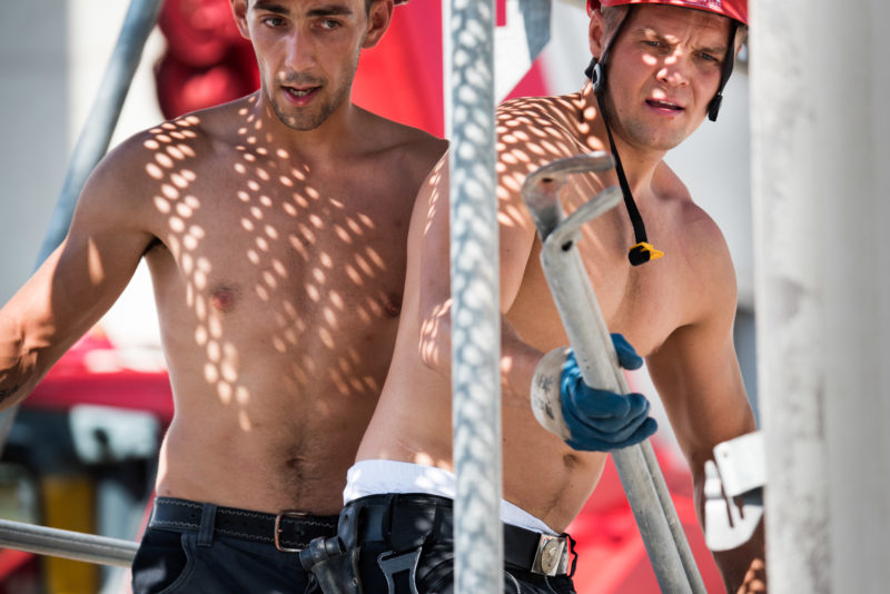 Editorial photography: Two scaffold workers work on one of the scaffold floors and attach props. The sun shines through the perforated plates onto their naked skin.