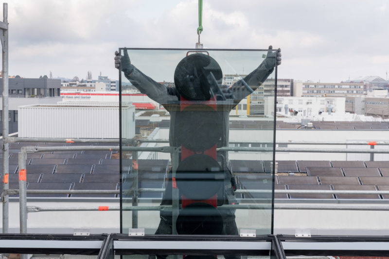 Reportagefotografie: Ein Fassadenbauer balanciert eine riesige Glasscheibe an ihre Position, die ihm am Kran hängend angeliefert wird.