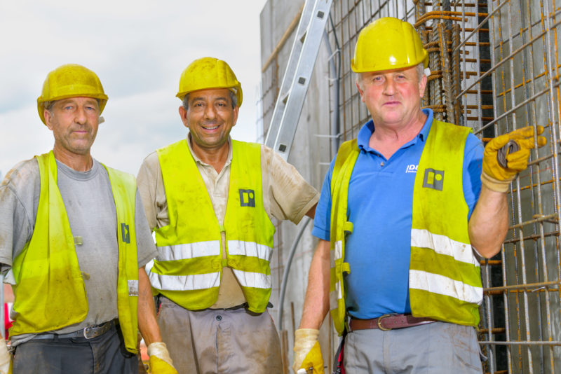 Editorial photography: Three construction workers in signal vests and yellow safety helmets proudly look into the camera. In the background you can see the reinforcing irons for a wall.