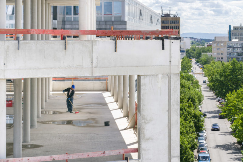 Reportagefotografie: Ein Arbeiter mit blauem Helm kehrt den Betonboden im noch fensterlosen Gebäude. Man sieht die Umgebung mit einer Straße mit Autos und grünen Bäumen.