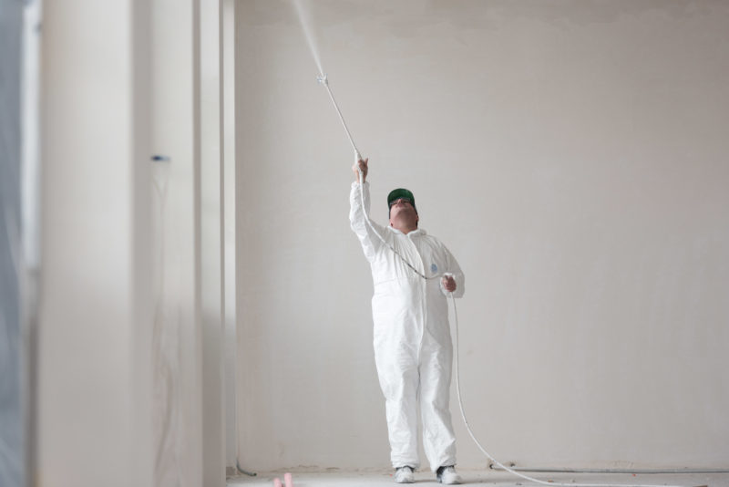 Editorial photography: A painter in white overalls sprays white paint on the ceiling in a white environment.