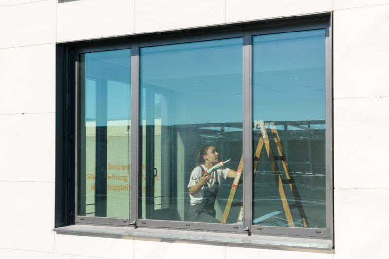 Editorial photography: A painter climbs the ladder to draw joints on the ceiling of an office. She is photographed from outside through a large window in which the blue sky is reflected.