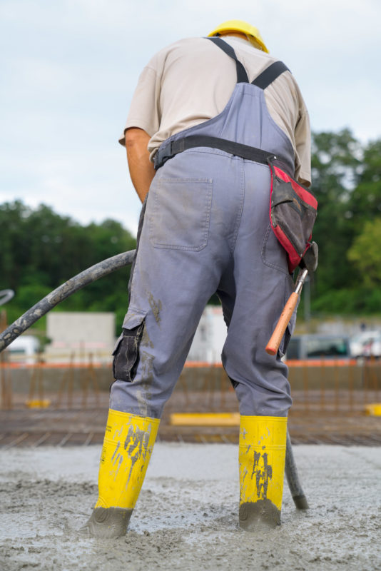 Reportagefotografie: Ein von hinten fotografierter Arbeiter steht mit gelben Gummistiefeln im noch flüssigen Beton einer Zwischendecke, um ihn mit einer Rüttelflache zu verdichten.