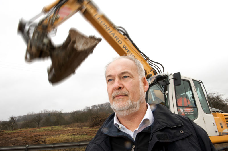 Mitarbeiterfotografie: Ein Bauleiter draußen auf einer Baustelle. Im Hintergrund sieht man einen großen Gelben Bagger, der gerade seine Schaufel ausfährt. Die Schaufel ist durch Bewegungsunschärfe dynamisch fotografiert.