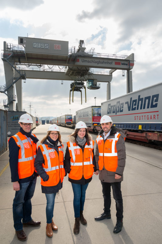 Group photo: Group picture of 4 visitors of a container station. In the background you can see a loading crane and freight car of the railway. All wear orange signal vests and white helmets.