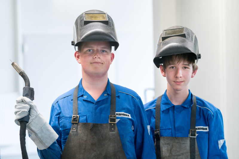 Editorial photography: Two pupils during a practical training at Liebherr-Hydraulikbagger GmbH. They practice welding for the first time. For that, they wear helmets with a sweat visor, thick aprons and leather gloves.