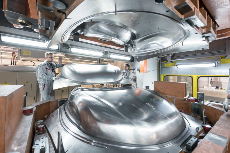 Industrial photography: Employees remove a completely pressed engine hood from a sheet metal press. The two halves of the mould shine metallically, while the finished hood shimmers rather matt.