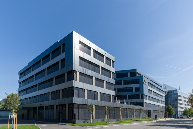 Architekturfotografie: Gesamtansicht eines neuen Bürogebäudes mit dunkler, in der Sonne glänzender Metallfassade. Der blaue Himmel sorgt für einen guten Kontrast der Architekturelemente.