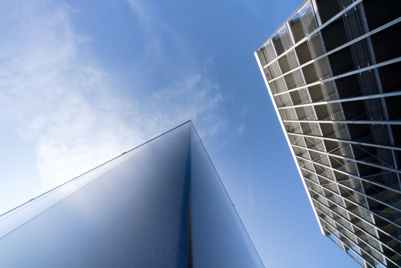 Architekturfotografie: Senkrecht gegen den leicht bewölkten blauen Himmel fotografiert, sieht man auf der einen Seite die glänzende Metallfassade eines Bürogebäudes und gegenüber die offene Struktur des angrenzenden Parkhauses.