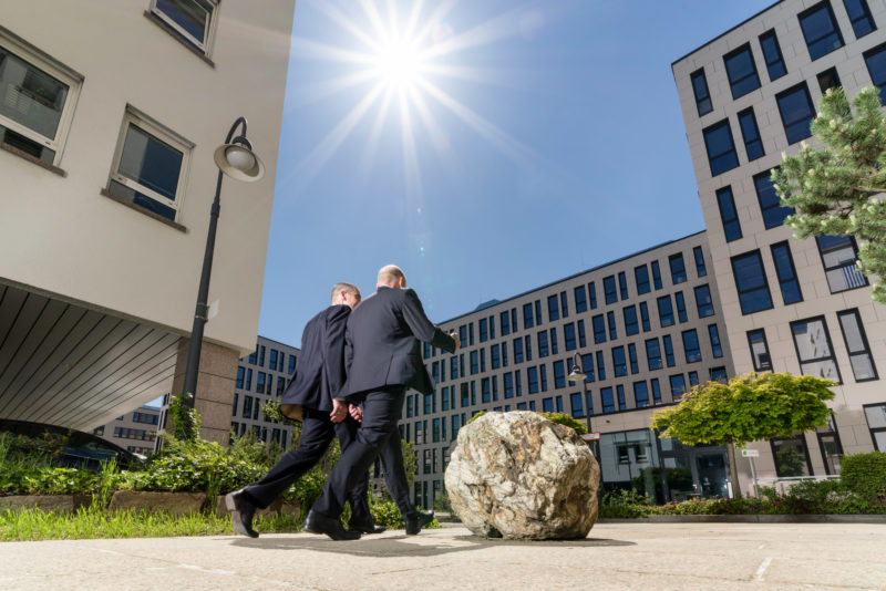 Architectural photography: Two employees leave their building in a modern office environment. You can see the sun against the blue sky between the towering companies in the area. In the foreground a round rock as decoration and behind it green trees and bushes.