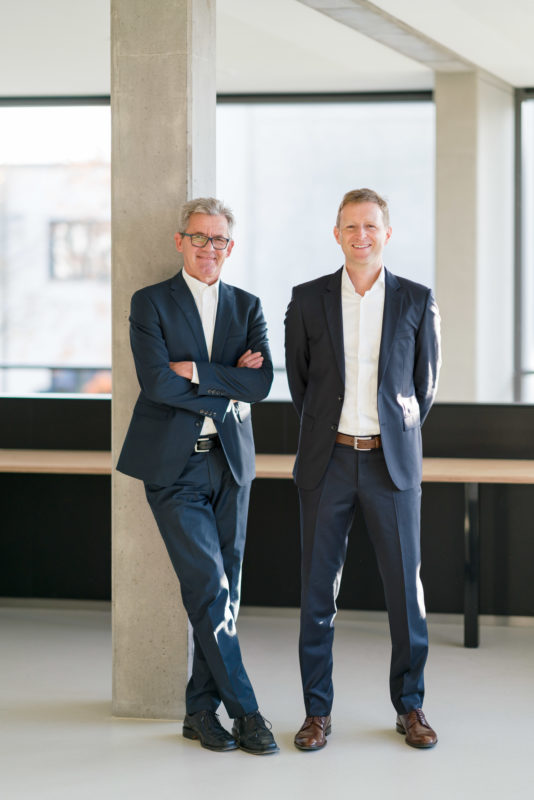 Managerportrait:  Two managers in suits with white shirts and no ties are standing laughing in the hallway of their company. The photo is a business portrait of managers and is suitable for annual reports and other representative company publications. It was photographed with a light telephoto lens in sunlight and with an open aperture. In addition, a flash system was used for brightening and better illumination.