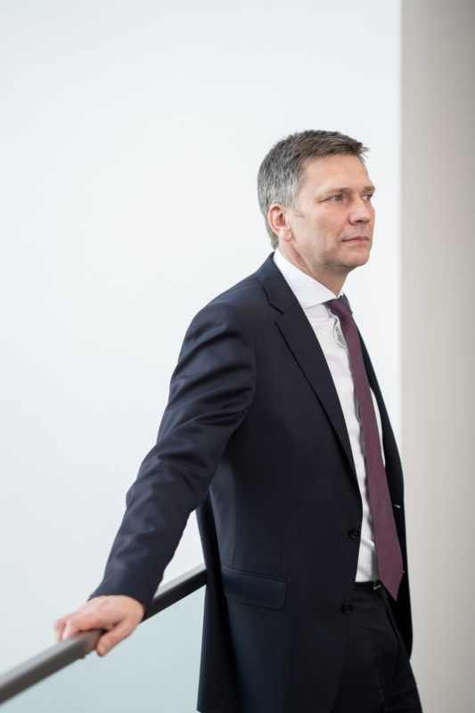 Portrait of a managing director: A male executive loosely leaning on a modern glass railing. He wears a tie and looks relaxed into the room.