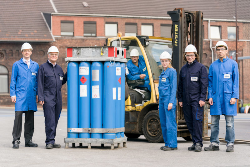 Gruppenfoto: Ganzkörperportrait von 6 Beschäftigten in der chemischen Produktion. Zwischen Ihnen stehen Gasbehälter. Einer sitzt in einem Gabelstapler.