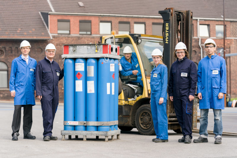 Group photo: Full body portrait of 6 employees in chemical production. There are gas tanks between them. One