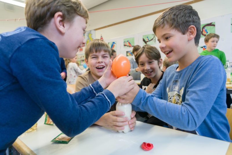 Reportagefotografie: Schüler haben bei einem einfachen chemischen Experiment viel Spaß. Sie mischen Essig und Backpulver in einer Flasche und sammeln das dabei entstehende Kohlendioxidgas in einem sich dann selbständig aufblasenden Luftballon.