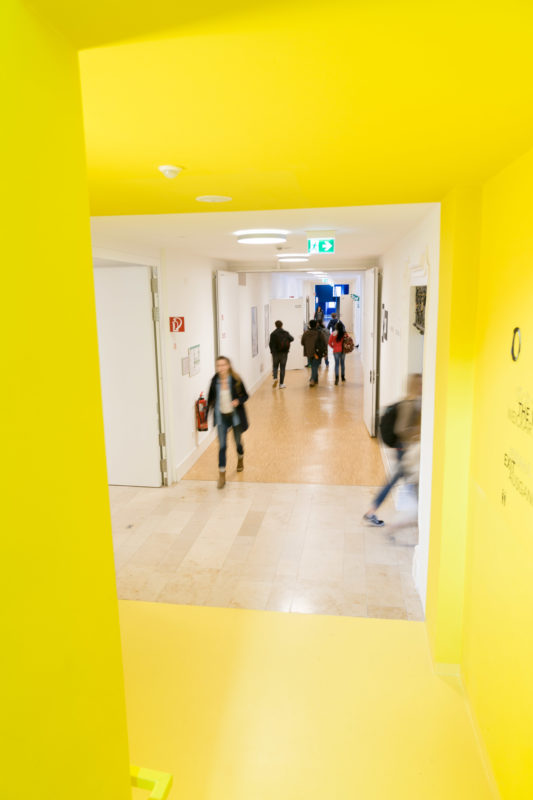 Architekturfotografie: Blick in den Gang eines privaten College, für das ein aufgegebenes Kloster umgebaut wurde. Durch ein gelbes Treppenhaus sieht man, wie Schüler in der Pause ihre Klassenräume wechseln.