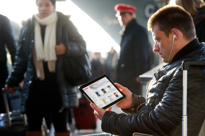 Reportage-Fotografie: Ein Mann sitzt am Bahnsteig und schaut auf sein Tablet-Computer. Im Ohr hat er Kopfhörer. Im Hintergrund steht ein Bahnmitarbeiter.