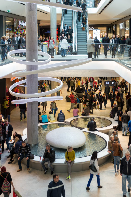 Architekturfotografie: Innenansicht vom Das Gerber, einem neuen Einkaufszentrum in der Stuttgarter Innenstadt im Gerberviertel. An gut besuchten Tagen bevölkern eine Menge Besucher das im Inneren mit einem Brunnen und einer Plastik geschmückte Gebäude.