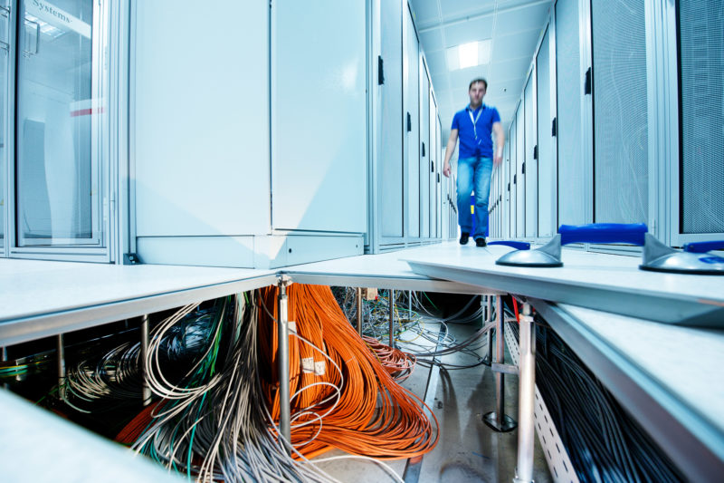 Industriefotografie: Blick in den geöffneten doppelten Boden eines Serverraumes. Man sieht eine unglaubliche Menge von Datenkabeln und Stromleitungen, während ein Mitarbeiter über die noch geschlossenen Platten des doppelten Bodens zwischen den Serverschränken hindurchgeht.