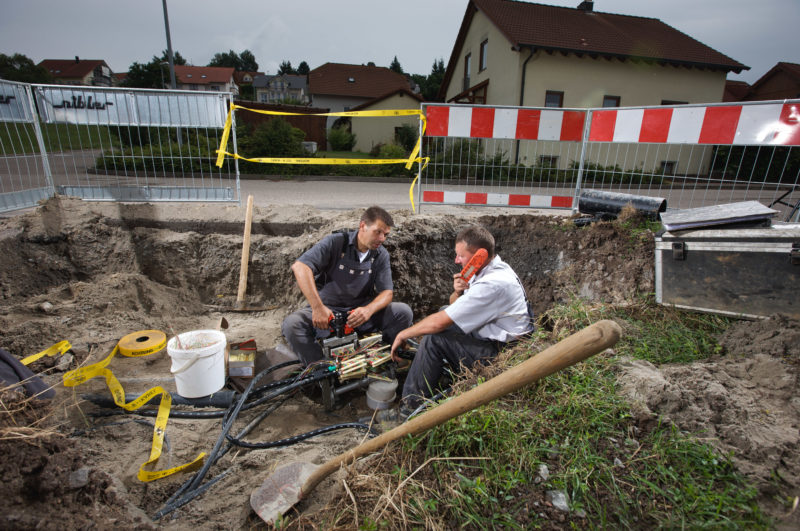 Industriefotografie: Zwei Techniker sitzen in einer Baugrube und arbeiten an Telefon- und Datenverbindungen für die Wohnsiedlung im Hintergrund.