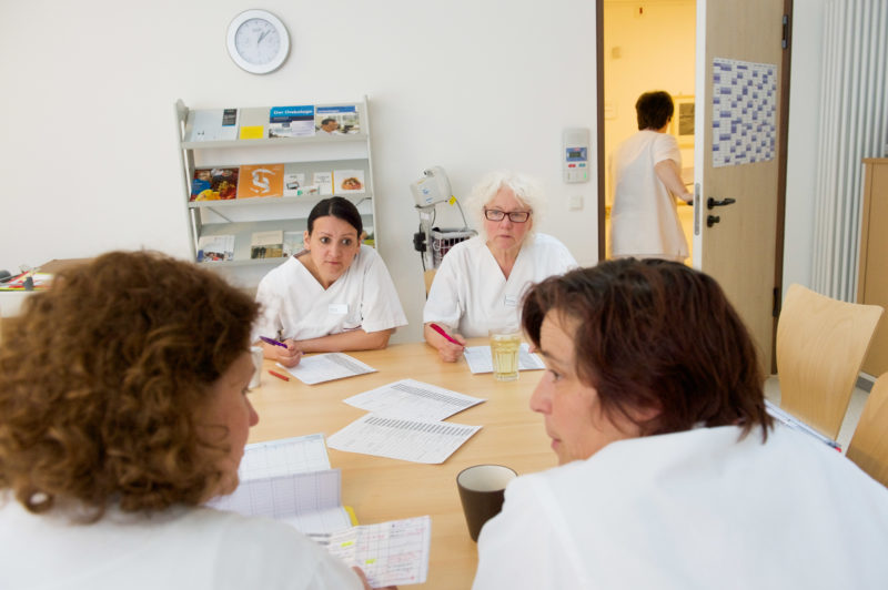 Medizinfotografie: Krankenpflegerinnen bei einer Besprechung im Dienstzimmer einer Station in einem Kreiskrankenhaus. Eine Schwester verlässt das Zimmer, um auf einen Patientenruf zu reagieren.