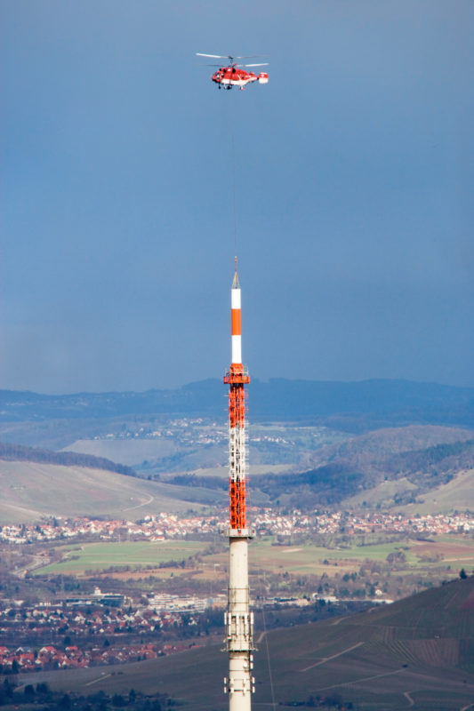 Technikfotografie: Ein Lastenhubschrauber hilft bei der Montage einer tonnenschweren DVB-T Sendeantenne an der Spitze eines Funkturms.