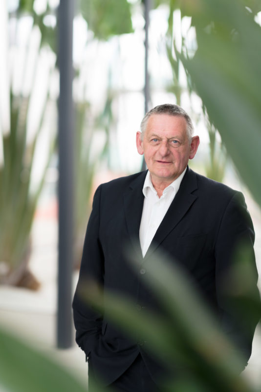 Managerportrait: The director of a research institute stands in the green inner courtyard of the building. The green covers his figure unsharply. Everything looks modern and bright. It is illuminated with a flash unit.