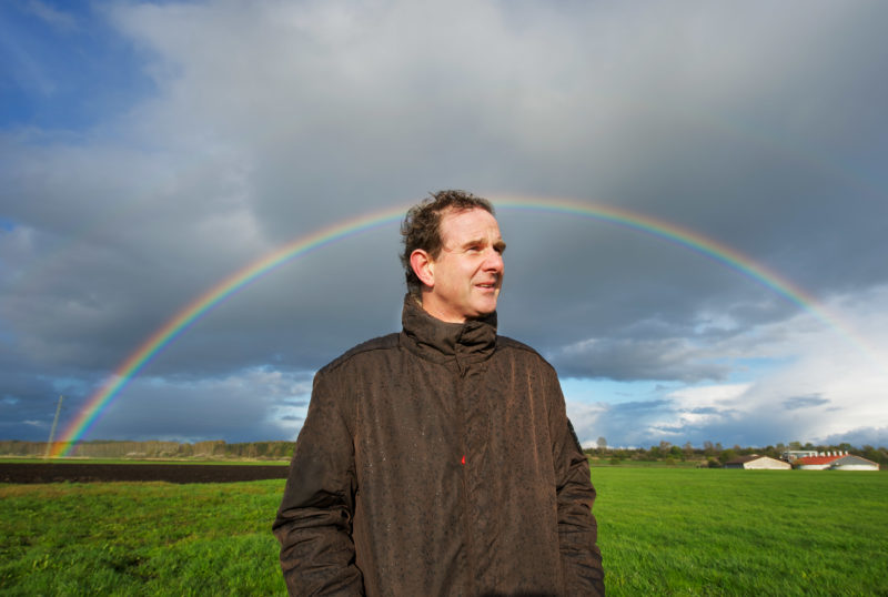 Mitarbeiterfotografie: Der Archäologe  Dirk Krauße steht auf einem Feld. Es regnet und vor den dunklen Wolken im Himmel glänzt ein Regenbogen.