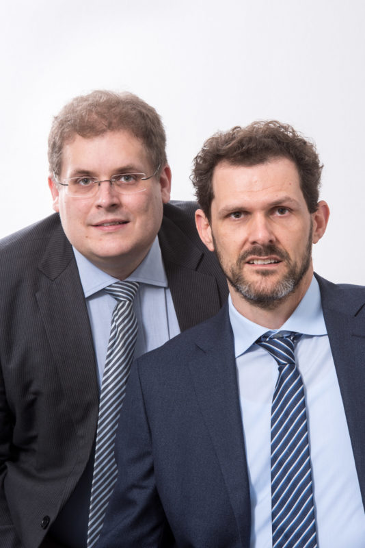 Managerportrait: Zwei Partner einer Firma im nahen Doppelportrait. Beide tragen blau gestreifte Krawatten. Der Hintergrund ist weiß.