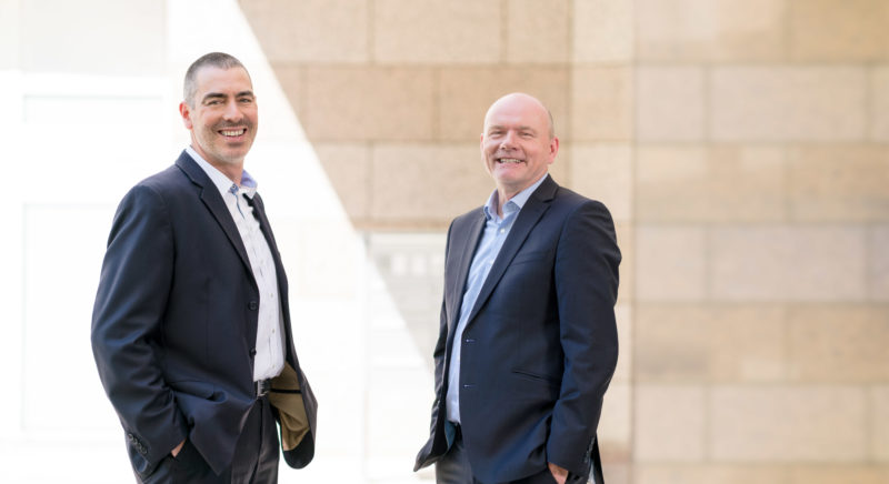 Managerportrait: Double portrait of two managing directors of a medium-sized company. They stand in the shade in front of the building and are illuminated with flash so that they stand out from the sunlit house wall made of large beige stone slabs.