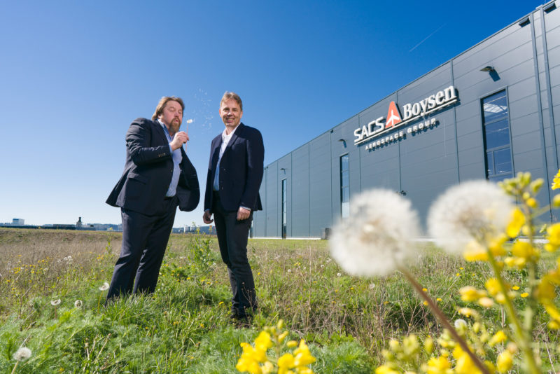 Managerportrait: Double portrait of two managers. They stand on a wild meadow in front of their modern company building, on which the logo of the company is attached. One has mature dandelion stems in his hand and blows the dandelion flowers to his colleague.