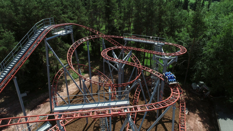 Aerial and Drone photography: A small roller coaster is tested with water-filled dummy passengers. The iron construction is surrounded by green forest.