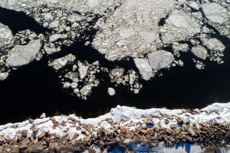 Luftaufnahmen und Drohnenfotografie: Senkrechte Drohnenaufnahme vom Eis auf dem Waser und dem Schnee am steil abfallenden steinigen Ufer an der finnischen Ostseeküste im Winter.