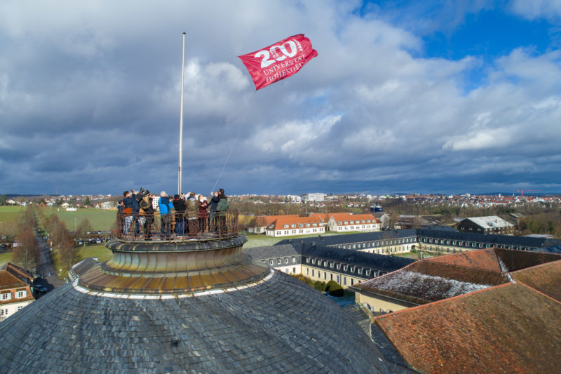 Luftaufnahmen und Drohnenfotografie: Auf dem Dach des Schlosses an der Universität Hohenheim wird anlässlich des 200 jährigen Jubiläums eine Fahne gehisst. Am dunkelblauen Himmel sind dramatische Wolken.